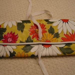 May include: A floral print clutch featuring large daisies in white, orange, yellow, and pink against a yellow background. The clutch has a white trim and a white ribbon closure. The overall theme is vintage and retro.