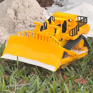 May include: A vibrant yellow toy bulldozer with black detailing and a large front blade. The toy is set on green grass, with sand and foliage in the background. The bulldozer features a detailed design, including a cab and tracks.