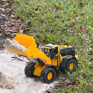 May include: A yellow and black toy front-end loader with the word "BOOTS" printed on the side. The loader's bucket is filled with sand. The toy is on a bed of sand and grass.
