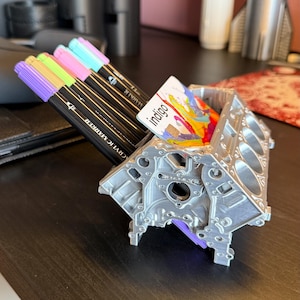 May include: A silver metal engine block desk organizer with a card holder. The card holder has a colorful card with the word "indigo" on it. The organizer is holding a set of colorful and black markers.