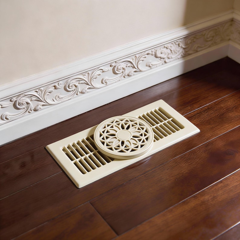 May include: An off-white rectangular floor vent with a decorative circular cover featuring a floral design. The vent is set on a dark brown hardwood floor, near a wall with ornate moulding.