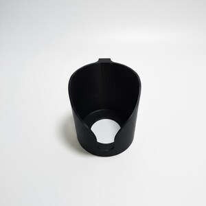 May include: A matte black cylindrical holder featuring a wide, open top and a circular cutout at the base. The object has a smooth, uniform texture and a small, protruding tab at the top rim, designed with a hollow interior and a solid, vertical wall structure.