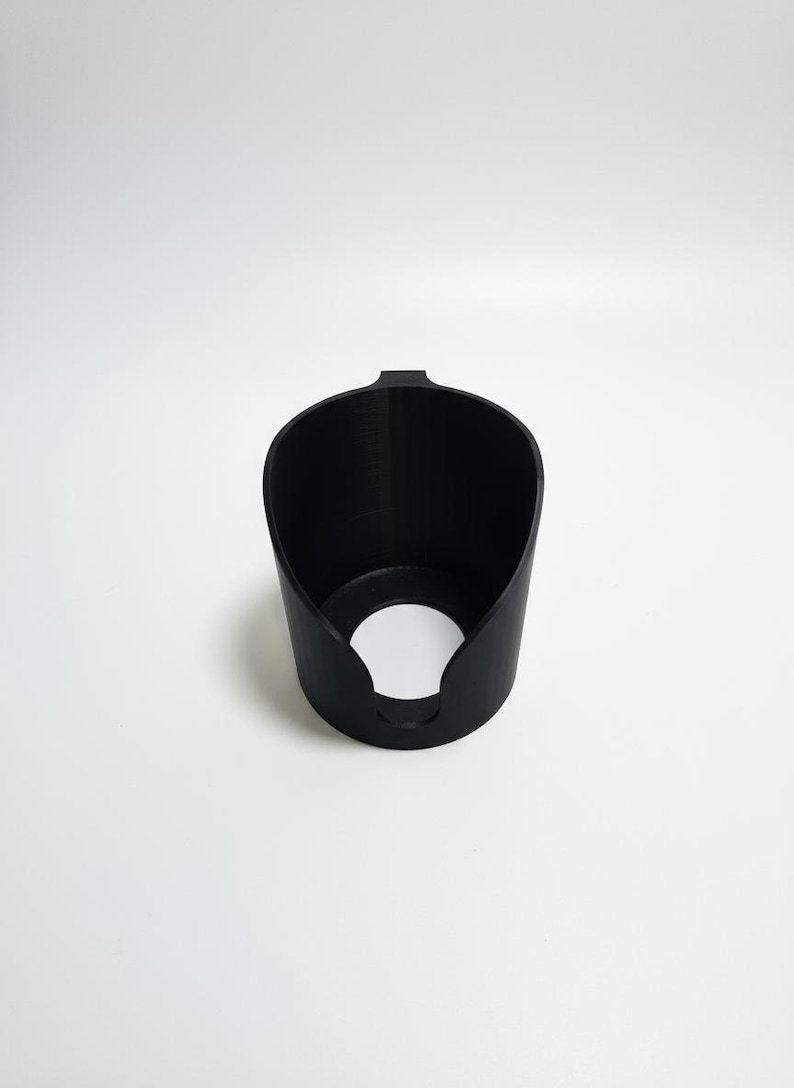 May include: A matte black cylindrical holder featuring a wide, open top and a circular cutout at the base. The object has a smooth, uniform texture and a small, protruding tab at the top rim, designed with a hollow interior and a solid, vertical wall structure.