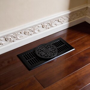 May include: A black metal floor vent with a decorative, circular cover featuring a floral design. The vent is rectangular with parallel slots. It's set into a dark wood floor, with a white wall and ornate trim in the background.