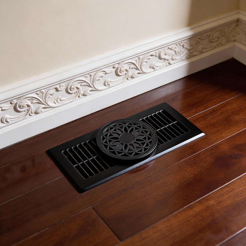 May include: A black metal floor vent with a decorative, circular cover featuring a floral design. The vent is rectangular with parallel slots. It's set into a dark wood floor, with a white wall and ornate trim in the background.