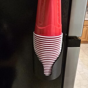 May include: A black plastic cup dispenser mounted vertically, holding a stack of red and white disposable plastic cups. The top red cup features the embossed text SOLO on its side, and the cups are stacked neatly within the curved holder.