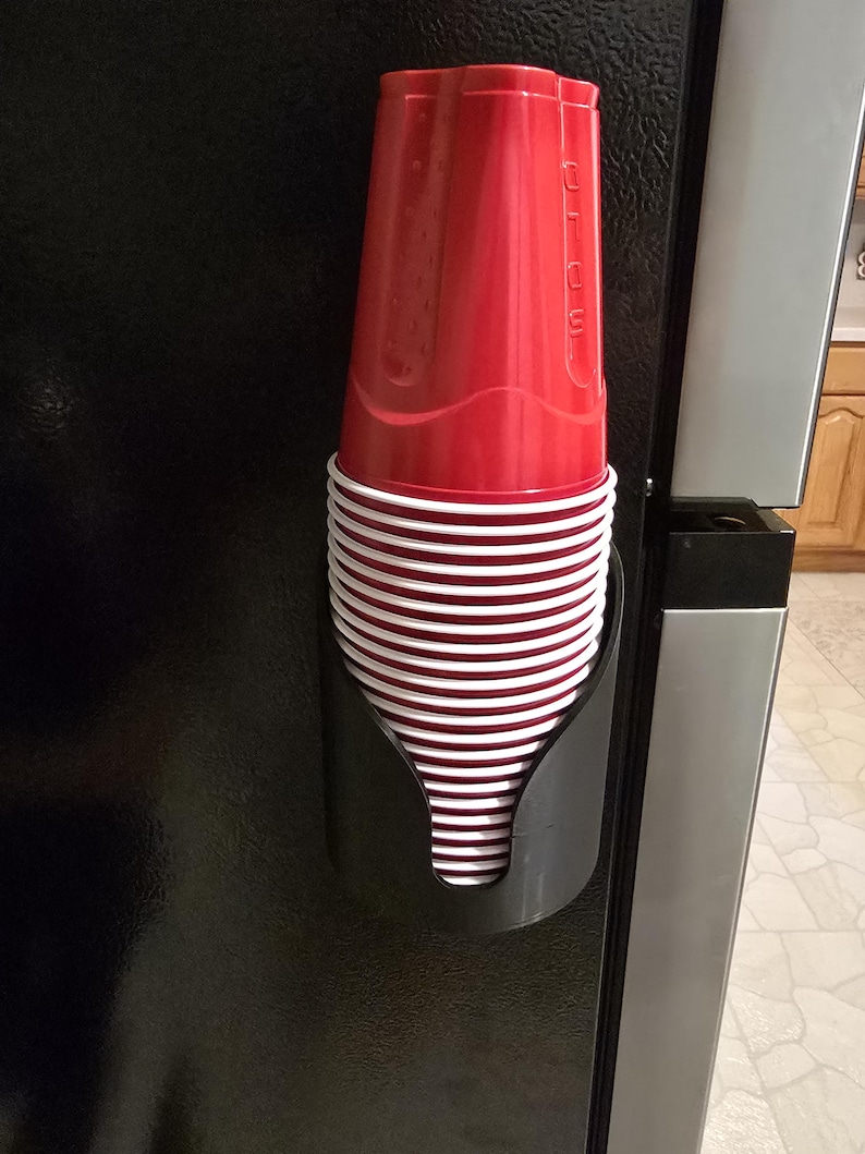 May include: A black plastic cup dispenser mounted vertically, holding a stack of red and white disposable plastic cups. The top red cup features the embossed text SOLO on its side, and the cups are stacked neatly within the curved holder.