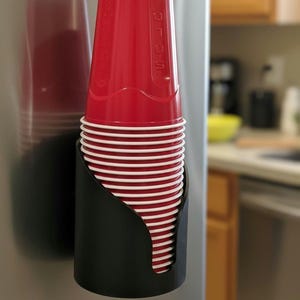 May include: A black plastic cup dispenser mounted vertically, holding a stack of red disposable plastic cups with white rims. The top cup features the embossed text Solo on its side. The dispenser is attached to a smooth, metallic surface.