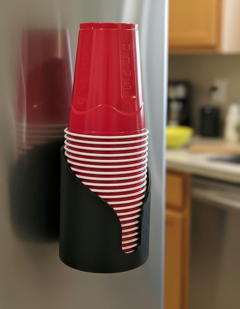 May include: A black plastic cup dispenser mounted vertically, holding a stack of red disposable plastic cups with white rims. The top cup features the embossed text Solo on its side. The dispenser is attached to a smooth, metallic surface.
