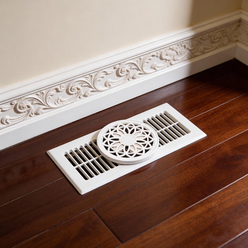 May include: A white floor vent with a decorative circular cover. The vent is set into a dark wood floor. The cover has a floral design. The background features a white wall and ornate trim.