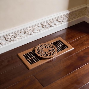 May include: A bronze floor vent with a decorative circular cover featuring a floral design. The vent is rectangular with parallel slots. The cover is placed on top of the vent. The vent is on a dark wood floor.