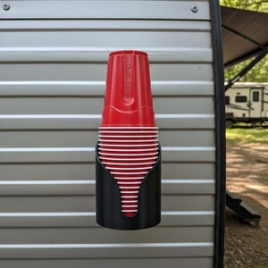 May include: A black plastic cup dispenser mounted to a wall, holding a stack of red plastic cups with white interior rims. The top cup features the embossed text SOLO. The dispenser has a curved, open-front design that secures the stacked cups.