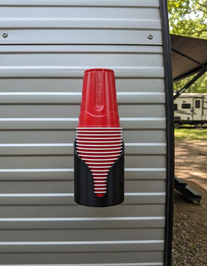 May include: A black plastic cup dispenser mounted to a wall, holding a stack of red plastic cups with white interior rims. The top cup features the embossed text SOLO. The dispenser has a curved, open-front design that secures the stacked cups.