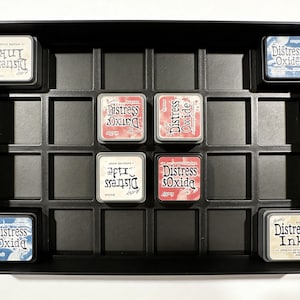 May include: A black ink storage tray filled with Distress Ink and Distress Oxide ink pads. The ink pads are square, with labels in various colors and fonts. The tray has multiple compartments to hold the ink pads.