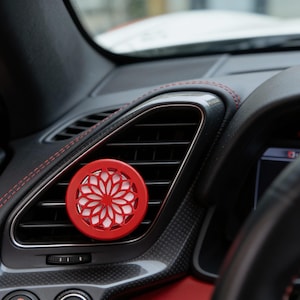 May include: A red car air vent diffuser with a floral design. The diffuser is circular and has a white interior. It is installed in a black car interior with red stitching.
