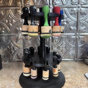 May include: A black, multi-tiered stamp holder with a circular base and multiple levels to hold stamps. The stamps have wooden handles and colorful tops in various colors, including black, green, red, and blue. The holder is designed to organize and display stamps.