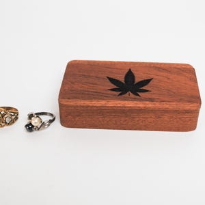 May include: A wooden box with a black marijuana leaf design on the lid. Two rings are in the foreground, one gold and one silver.