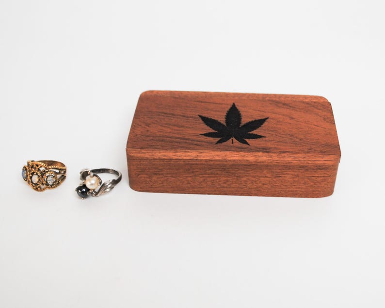 May include: A wooden box with a black marijuana leaf design on the lid. Two rings are in the foreground, one gold and one silver.