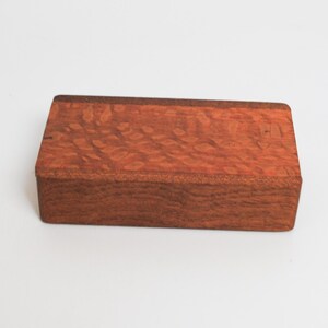 May include: A rectangular wooden box with a reddish-brown color and a wood grain pattern. The box has a smooth surface and appears to be made of high-quality wood.