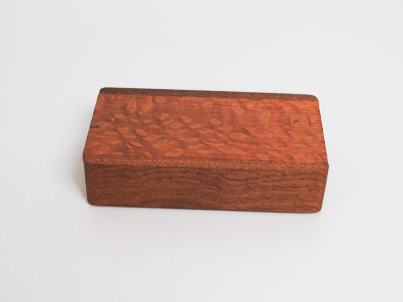 May include: A rectangular wooden box with a reddish-brown color and a wood grain pattern. The box has a smooth surface and appears to be made of high-quality wood.
