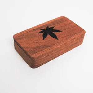 May include: A small wooden box with a dark brown finish. The top of the box has a black leaf design.