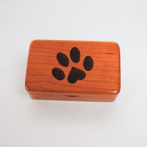 May include: A small wooden box with a paw print design on the lid. The box is made of cherry wood and has a natural finish.