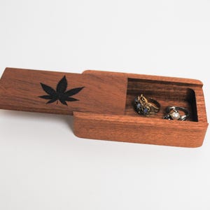 May include: A wooden ring box with a hinged lid. The lid has a black leaf design. The box is open and two rings are inside.