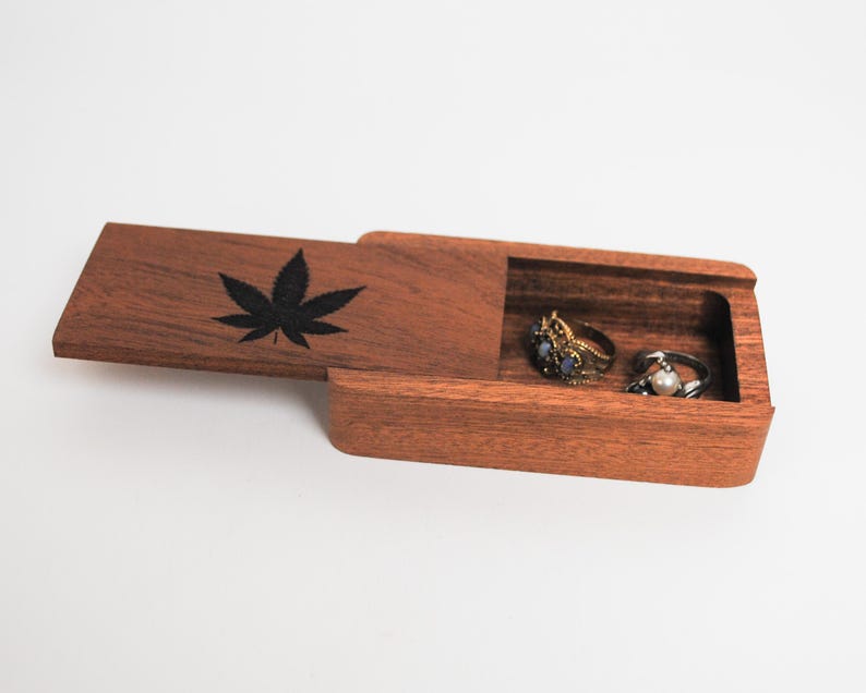 May include: A wooden ring box with a hinged lid. The lid has a black leaf design. The box is open and two rings are inside.