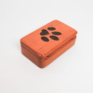 May include: A small wooden box with a hinged lid. The lid is decorated with a paw print and a heart. The box is made of cherry wood and has a natural finish.