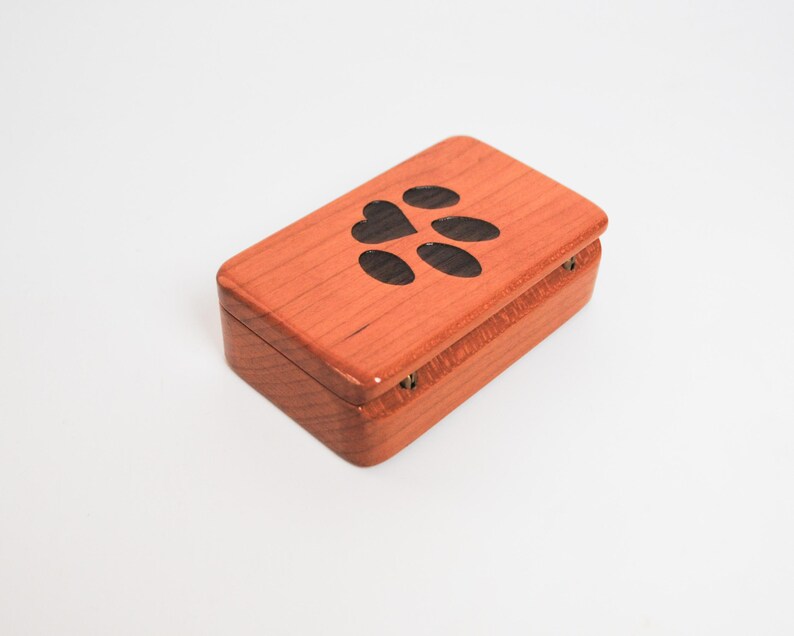 May include: A small wooden box with a hinged lid. The lid is decorated with a paw print and a heart. The box is made of cherry wood and has a natural finish.
