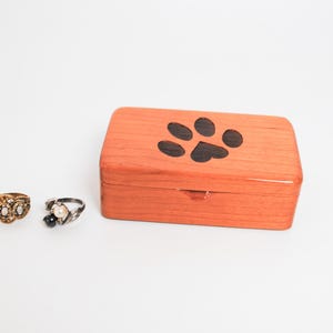 May include: A small wooden box with a paw print design on the lid. The box is made of light wood and has a natural finish. The paw print is black and is centred on the lid. The box is closed and is sitting on a white surface.