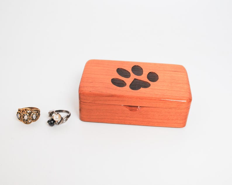 May include: A small wooden box with a paw print design on the lid. The box is made of light wood and has a natural finish. The paw print is black and is centred on the lid. The box is closed and is sitting on a white surface.