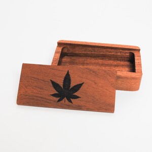 May include: A wooden box with a hinged lid. The lid has a black marijuana leaf design engraved on it. The box is made of dark wood and has a natural finish.