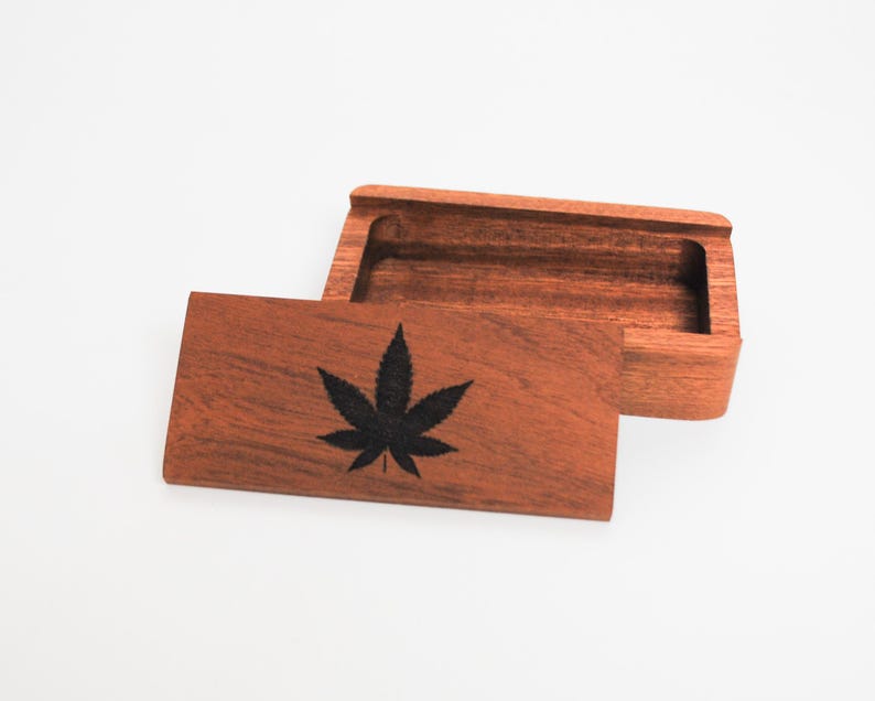 May include: A wooden box with a hinged lid. The lid has a black marijuana leaf design engraved on it. The box is made of dark wood and has a natural finish.