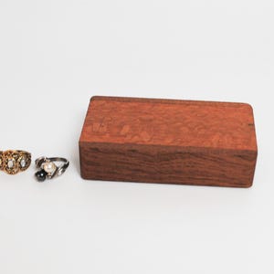 May include: Two rings on a white surface. The ring on the left is gold with three white stones. The ring on the right is silver with a black stone and a white stone. A small wooden box is in the background.