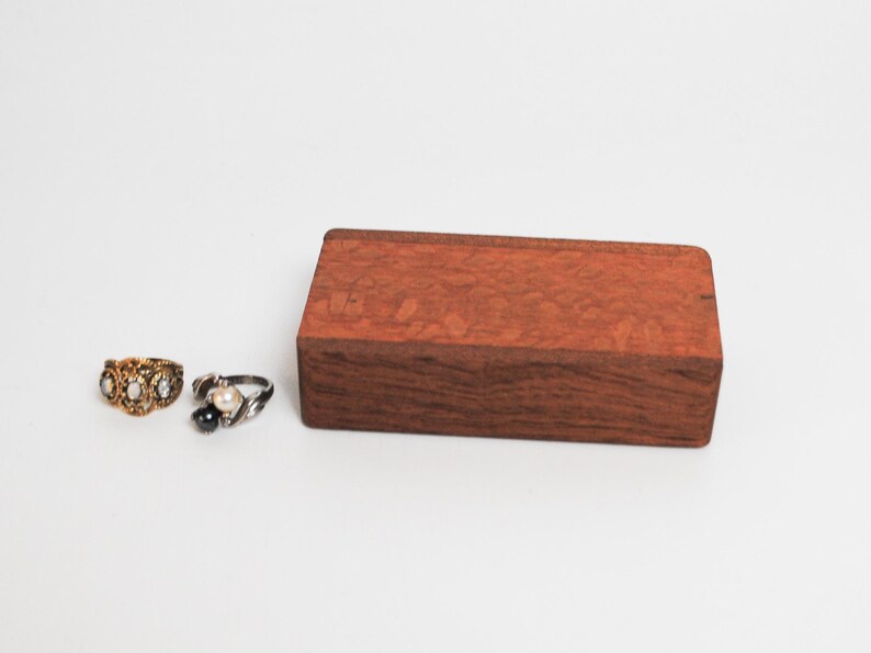 May include: Two rings on a white surface. The ring on the left is gold with three white stones. The ring on the right is silver with a black stone and a white stone. A small wooden box is in the background.