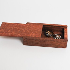 May include: A small wooden box with a hinged lid, open to reveal three rings inside. The box is made of dark wood with a reddish-brown hue and a patterned grain.