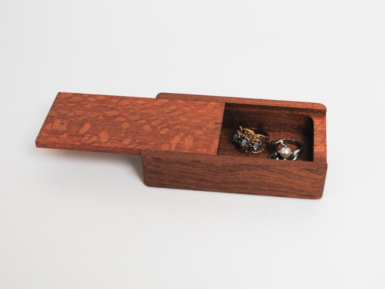 May include: A small wooden box with a hinged lid, open to reveal three rings inside. The box is made of dark wood with a reddish-brown hue and a patterned grain.