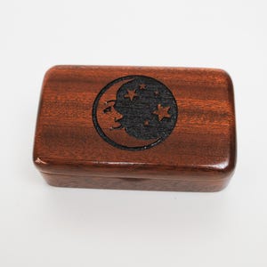 May include: A rectangular wooden box with a dark brown finish. The lid features a black circular design with a crescent moon and stars. The box is likely used for storing small items, such as jewellery or keepsakes.