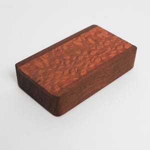 May include: A small wooden box with a red and brown wood grain pattern on the top. The box is made of two different types of wood.
