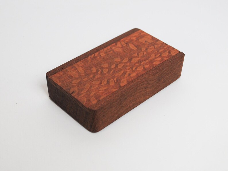 May include: A small wooden box with a red and brown wood grain pattern on the top. The box is made of two different types of wood.