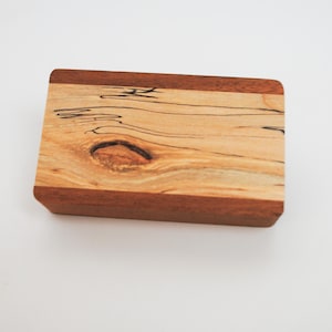 May include: A small wooden box with a natural wood finish and a dark brown border. The box has a knot in the wood and a unique grain pattern.
