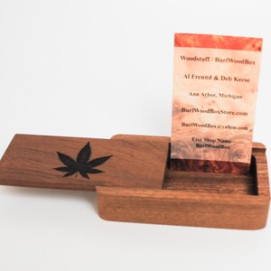 May include: A wooden box with a hinged lid, featuring a black leaf design on the lid. The box is open, revealing a business card inside. The card has a wood grain background and text that reads "Woodstuff / BurlWoodBox", "Al Freund & Deb Keese", "Ann Arbor, Michigan", "BurlWoodBoxStore.com", "BurlWoodBox@yahoo.com", "Etsy Shop Name - BurlWoodBox".