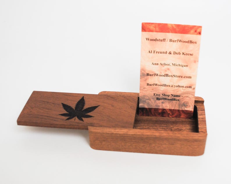 May include: A wooden box with a hinged lid, featuring a black leaf design on the lid. The box is open, revealing a business card inside. The card has a wood grain background and text that reads "Woodstuff / BurlWoodBox", "Al Freund & Deb Keese", "Ann Arbor, Michigan", "BurlWoodBoxStore.com", "BurlWoodBox@yahoo.com", "Etsy Shop Name - BurlWoodBox".