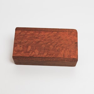 May include: A small wooden box with a reddish-brown finish. The box has a natural wood grain pattern and a smooth surface.