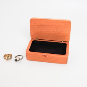 May include: A small wooden box with a black velvet interior. The box is open and two rings are sitting on a white surface in front of it.