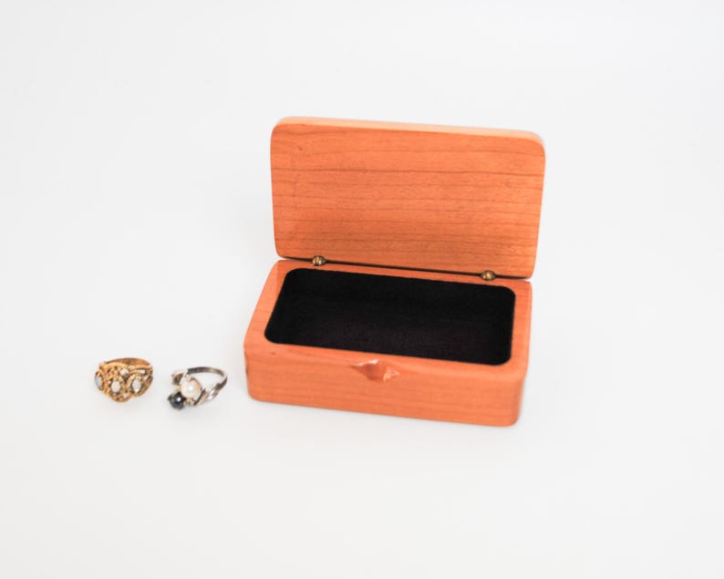 May include: A small wooden box with a black velvet interior. The box is open and two rings are sitting on a white surface in front of it.