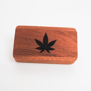 May include: A small wooden box with a dark brown finish. The box has a black marijuana leaf design on the top.