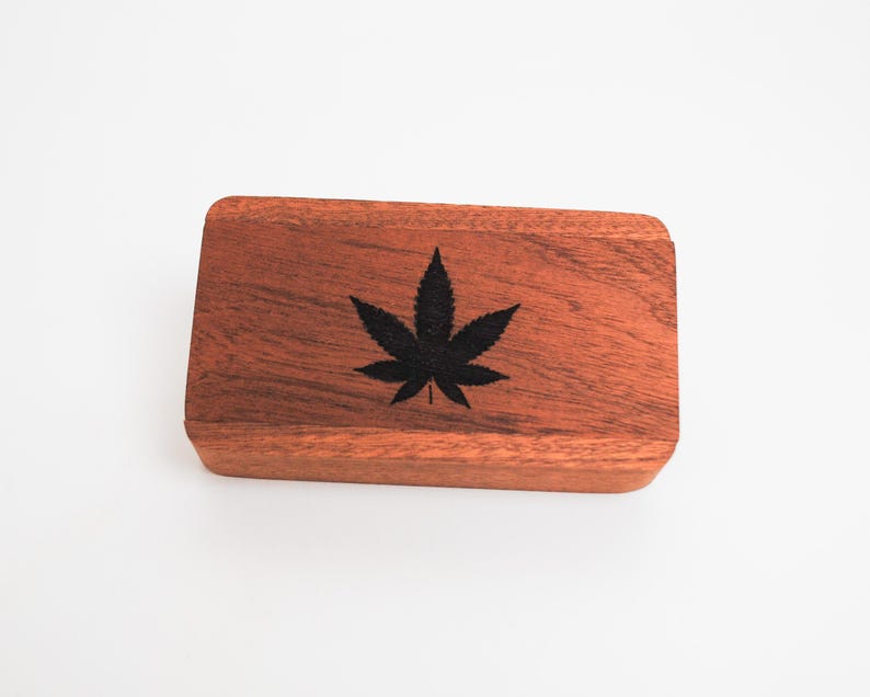 May include: A small wooden box with a dark brown finish. The box has a black marijuana leaf design on the top.