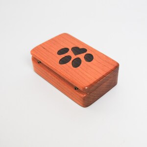 May include: A small wooden box with a hinged lid. The lid features a paw print design with a heart in the centre. The box is made of light brown wood.